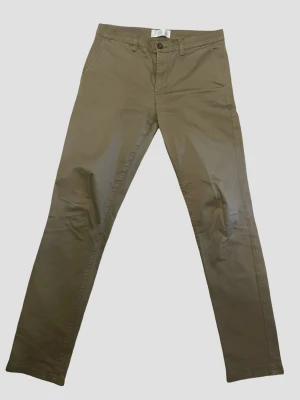 Beiga chinos från These Glory Days - Snygga beiga chinos från These Glory Days i storlek 34 (50) med klassisk rak passform och diskreta fickor både fram och bak. Tillverkade i mjukt bomullstyg som ger en clean look. Perfekta för dig som gillar stilrena och enkla byxor.