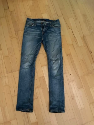 Blå nudie jeans - Säljer ett par nudie jeans med slitningar och en liten reva på ena benet, tillverkade i denim med lätt stretch. Perfekta för en avslappnad och trendig look. Andvänts max 5 gånger. W31 L32