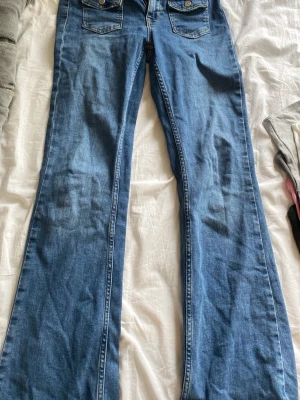 Blå bootcut jeans från Kappahl - Snygga blå bootcut jeans från Kappahl med klassiska fickor fram och bak. Jeansen har kontrastsömmar och en cool knappdetalj vid gylfen. Perfekt för dig som gillar en avslappnad och trendig stil. Materialet är denim.