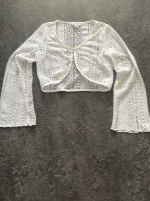 Vit virkad bolero med vida ärmar - Säljer en vit, virkad bolero med snyggt mönster och vida ärmar. Kort modell som passar perfekt över linnen eller klänningar. Tröjan har öppen front och är gjord i ett lätt och luftigt material.