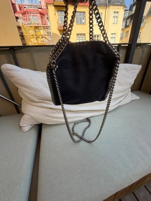 Svart Stella McCartney handväska - Snygg blå handväska från Stella McCartney med kedjedetaljer i silver runt kanterna och som axelrem. Väskan har en mjuk, vegansk mockaliknande yta och magnetlås. Märkt 'Made in Italy' på insidan. Perfekt för dig som gillar trendiga accessoarer.