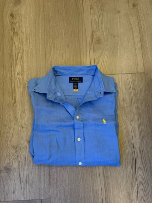 Blå linne skjorta från Polo Ralph Lauren - Klassisk blå linne skjorta från Polo Ralph Lauren med krage och vita knappar. Skjortan har lång ärm och den ikoniska gula logotypen broderad på bröstet. Det är en linne skjorta som är perfekt för sommaren☀️Det är storlek L i barnstorlek men jag skulle säga att den passar XS! Skriv vid minsta fundering!✅