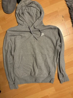 Grå hoodie från Nike med logga - Klassisk grå hoodie från Nike med vit broderad logga på bröstet. Hoodien har huva med dragsko och en stor magficka framtill. Tillverkad i mjukt sweatshirtmaterial som är perfekt för chill dagar. Ribbade muddar vid ärmslut och nederkant.
