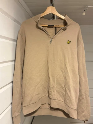 Beige halfzip från Lyle & Scott - Snygg beige halfzip-tröja från Lyle & Scott med klassisk gul logga på bröstet. Tröjan har hög krage med dragkedja och långa ärmar. Perfekt för en avslappnad och stilren look.