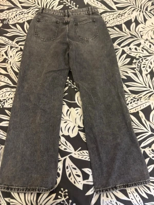 Gråa raka jeans med slitningar - Säljer ett par gråa jeans med raka ben och stora slitningar på båda knäna. Jeansen har klassisk femficksmodell och råa kanter vid bensluten. Perfekta för en avslappnad och trendig look.