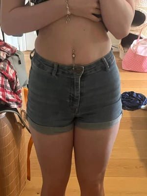 Blå denimshorts med hög midja - Säljer ett par klassiska blå denimshorts med hög midja och uppvikta benslut. Shortsen har fem fickor och stängs med dragkedja och knapp. Perfekta för varma dagar och ger en snygg, avslappnad look. Använda mycket och ända synliga slitaget är lite lite vid bakfickorna men inget man tänker på, annars helt som nya. 