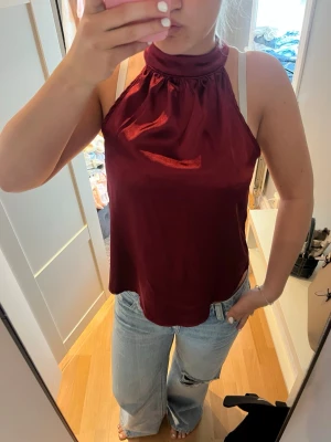 Vinröd halterneck linnetopp - Snygg vinröd halterneck linnetopp i glansigt material med hög krage. Toppen är ärmlös och har en lös passform, perfekt att styla med jeans eller kjol. Ger en trendig och chic look.