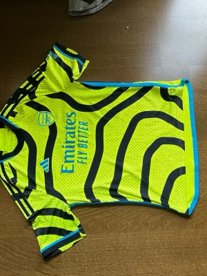 Arsenal tröja Adidas gul - Snygg Arsenal tröja från Adidas i neon-gult med svarta och blå detaljer. Tröjan har korta ärmar, rund hals och coolt mönster över hela plagget. Tillverkad i lätt och ventilerande material, perfekt för fotbollsträning eller match.
