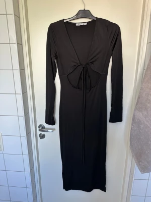 Svart långklänning från NAKD S - Svart långklänning från NAKD med djup v-ringning i ryggen och snygg snörningsdetalj. Klänningen har långa ärmar och är figurnära i modellen. Tillverkad i ett mjukt och stretchigt material som ger en bekväm passform.