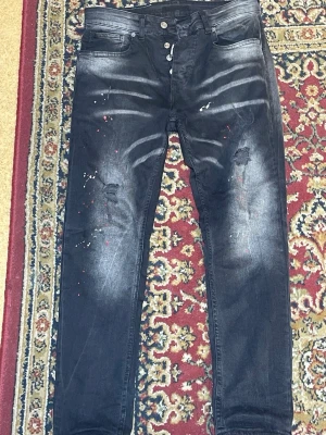 Svarta jeans ICON2 med slitningar - Säljer ett par svarta jeans från ICON2 med slitningar och färgstänk på framsidan. Jeansen har en tvättad look, fem fickor och knappgylf. Materialet är denim och de har en modern, avslappnad passform.