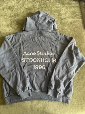 hoodie från Acne Studios - Snygg hoodie från Acne Studios med huva och ribbade muddar. På ryggen står det 'Acne Studios STOCKHOLM 1996' i vitt tryck. Perfekt oversized fit och mjukt material, troligen bomull. Enkel och clean framtill, statement-tryck bak.