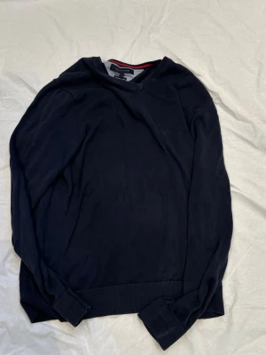 Mörkblå tröja Tommy Hilfiger - Klassisk mörkblå tröja från Tommy Hilfiger i premium bomull. Tröjan har rund halsringning med en snygg detalj i grått och rött vid kragen samt ribbade muddar vid ärmslut och nederkant. Perfekt för en stilren och enkel look.
