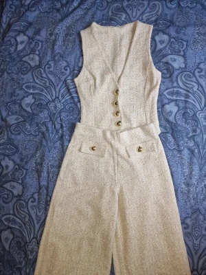 Viral boucléset med väst och vida byxor från Zara - Ett slutsålt viral set i beige bouclétyg med matchande väst och vida byxor. Västen är ärmlös med djup V-ringning och fyra guldknappar framtill. Byxorna har hög midja och dekorativa fickor. Perfekt för sommaren.