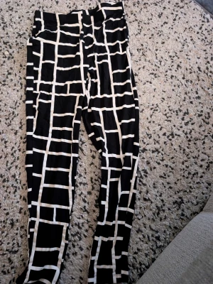 Svarta leggings med vit rutmönster stl L - Säljer ett par svarta leggings med vitt rutmönster. De har hög midja och är stretchiga för en bekväm passform. Materialet känns mjukt och följsamt, perfekt för dig som gillar grafiska mönster och vill sticka ut lite extra.