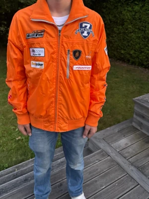 Orange racing bomberjacka Capenorth - Snygg orange bomberjacka från Capenorth med racing-inspirerade patches och broderier. Jackan har dragkedja framtill, muddar vid ärmslut och nederkant samt flera coola loggor och siffran 7 på ryggen. Materialet är glansigt och lätt, perfekt för dig som gillar sportig stil.