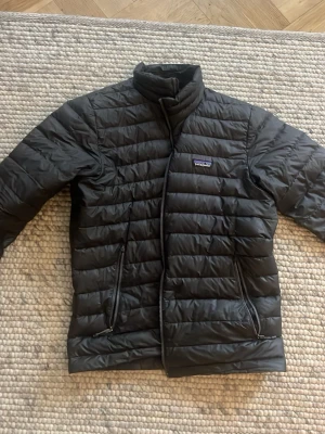 Svart pufferjacka från Patagonia - Säljer en svart pufferjacka från Patagonia med quiltad design och hög krage. Jackan har två dragkedjeförsedda fickor framtill och diskret logga på bröstet. Perfekt för kalla dagar och riktigt skön att bära. Lite lite sliten där bak men det kan man fixa gratis på Patagonia butiken 