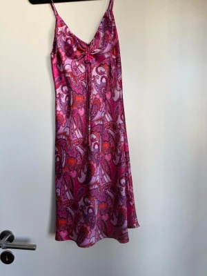 Rosa paisleymönstrat kläder  - Säljer en kläder med tunna axelband och v-ringning. Tyget är glansigt och har ett färgstarkt paisleymönster i rosa, lila och röda toner. Lätt och luftigt material som känns skönt mot huden.