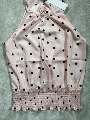 Ljusrosa prickig topp från Neo Noir - Supersöt ljusrosa topp från Neo Noir med bruna prickar. Toppen har smockad nederkant med volang och är ärmlös med halterneck. Tillverkad i ett glansigt, mjukt material som känns lyxigt och somrigt.