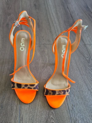 Neonorange pumps med leoparddetalj - Snygga neonorange pumps från EGO med smala remmar och leopardmönstrad detalj framtill. Skorna har öppen tå, smal klack och justerbar rem runt vristen. Perfekta för dig som vill sticka ut med färg och mönster. 12cm klack.