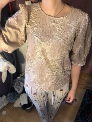 Beige strukturerad blus med puffärm - Säljer en beige blus med unik struktur och puffiga trekvartsärmar. Blusen har rund halsringning och ett lätt glansigt tyg som ger en lyxig känsla. Passar perfekt till både jeans och kjol för en trendig look.