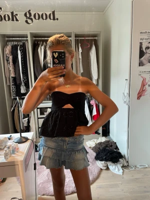 Svart bandeau topp med broderi - Säljer en svart bandeau topp med broderade detaljer och en liten cut-out framtill. Toppen är axelbandslös och har en lätt utsvängd passform nedtill. Perfekt för dig som vill ha en trendig och luftig topp till sommaren.