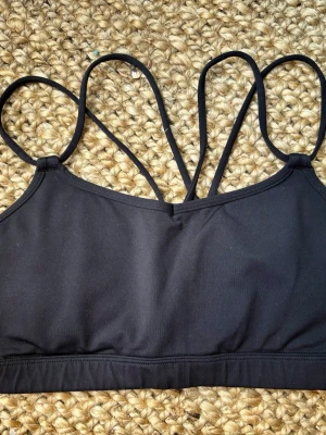 Lululemon Svart sport-bh med tunna axelband - Snygg svart sport-bh från Lululemon med flera tunna korsade axelband i ryggen. Bh:n är i stretchigt material som känns mjukt mot huden och ger bra stöd. Vadderade kupor. Perfekt för träning eller chill. 