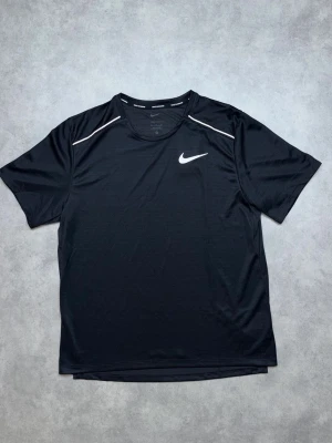 Svart Nike tränings-tshirt - Snygg svart tränings-tshirt från Nike med vit logga på bröstet och vita detaljer vid axlarna. Tillverkad i ett lätt och snabbtorkande funktionsmaterial som passar perfekt till träning och sportaktiviteter. Klassisk rund hals och korta ärmar.