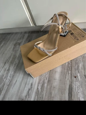 Beige sandalett med strassdetaljer - Trendig sandalett i beige med fyrkantig tå och smala remmar dekorerade med glittrande strass. Klack i genomskinlig akryl ger en cool touch. Perfekt för dig som vill sticka ut med stilrena detaljer och modern design.