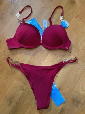 Vinröd bikini från Censored med gulddetaljer push up - Snygg vinröd bikini från Censored med vadderad bygel-bh och matchande trosa. Båda delarna har guldfärgade metallringar som detaljer på axelband och sidor. Perfekt för stranden eller poolen, med justerbara axelband för bästa passform. 