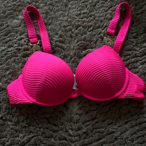 Rosa push-up bikiniöverdel med gulddetaljer  - Snygg rosa bikiniöverdel med vadderade kupor och guldfärgade detaljer på axelbanden. Bikinin har ribbad struktur och justerbara axelband för bästa passform. Perfekt för strand eller pool!