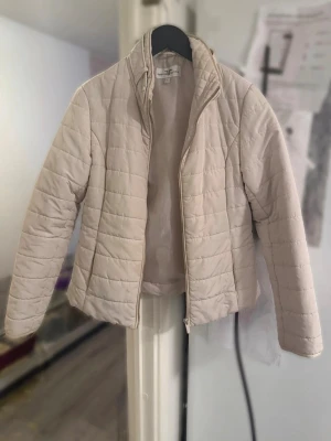 Beige quiltad jacka S - Stilren tunn pufferjacka med quiltad design och dragkedja framtill. Jackan har hög krage och långa ärmar, perfekt för kyligare dagar. Storlek S, använt men fint skick!
