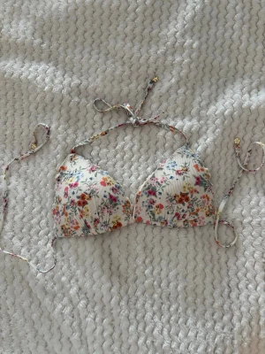 Blommig bikiniöverdel med knyt - Söt bikiniöverdel i trekantsmodell med blommigt mönster i flera färger på vit botten. Knyt i nacken och ryggen för justerbar passform. Perfekt för lata dagar på stranden eller poolen.