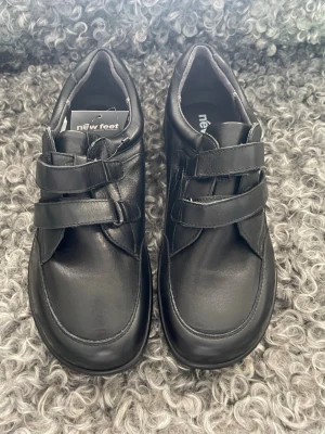 Svarta loafers med kardborreband - Snygga svarta loafers från New Feet i slätt läder med två breda kardborreband för enkel på- och avtagning. Skorna har en rundad tå och diskret söm längs kanterna. Perfekta för dig som gillar stilrena och bekväma skor.