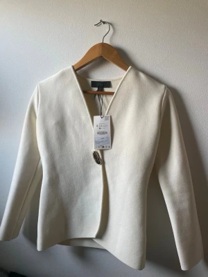 Vit kavaj med guldknapp från Zara - Stilren vit kavaj från Zara med unik asymmetrisk knäppning och en stor dekorativ guldknapp framtill. Kavajen har lång ärm, v-ringad hals och är tillverkad i ett strukturerat tyg som ger en exklusiv känsla. Perfekt för dig som gillar minimalistisk och modern stil.