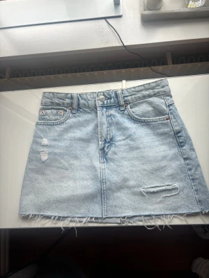 Ljusblå jeanskjol från Stradivarius - Trendig ljusblå jeanskjol från Stradivarius i klassisk femficksmodell. Kjolen har slitna detaljer, rå kant nertill och dragkedja med knapp framtill. Perfekt för dig som gillar en avslappnad och cool stil.