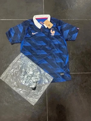 Blå Frankrike Nike fotbollströja - Snygg blå fotbollströja från Nike med Frankrikes landslagsmärke på bröstet. Tröjan har ett mönster i olika blå nyanser, vit krage och korta ärmar. Perfekt för dig som älskar fotboll och vill stötta Frankrike på match eller träning.