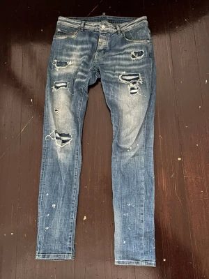 Blå slitna skinny Horspist jeans - Säljer ett par blå skinny jeans med slitningar och stora hål på framsidan. Jeansen har en tvättad look med ljusa partier och färgstänk för en trendig, avslappnad stil. Klassisk femficksmodell med knappgylf och bälteshällor. Perfekta för dig som gillar streetwear.