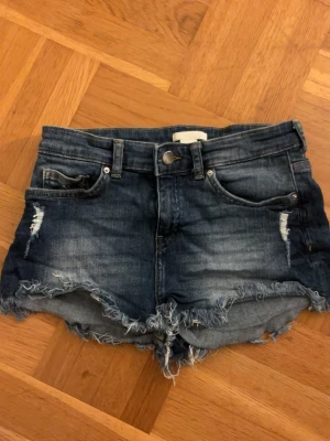 Blå jeansshorts - Säljer ett par blå jeansshorts med fransig, rå kant och slitna detaljer framtill. Shortsen har 4 fickor, bälteshällor och knappgylf. Perfekta för sommaren och ger en avslappnad vibe.