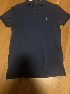 Ralph lauren - Säljer en klassisk marinblå polotopp från Polo Ralph Lauren med korta ärmar och krage med knappar. Ikoniska loggan broderad på bröstet. Perfekt för dig som gillar stilrena och tidlösa plagg.