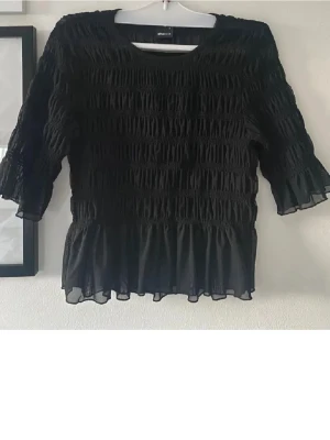 Gina Tricot - Supersöt svart blus från Gina Tricot med smockad struktur över hela toppen och volangkant nedtill. Perfekt till vardags eller fest🍾 Prioriterar snabb affär!
