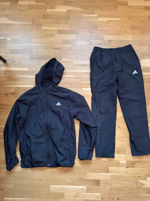 Svart träningsset från Adidas - Säljer ett svart träningsset från Adidas med jacka och byxor. Jackan har huva, dragkedja och Adidas-logga på bröstet. Byxorna har resår i midjan och logga på benet. Perfekt för träning eller uppvärmning. Materialet är lätt och andas.
