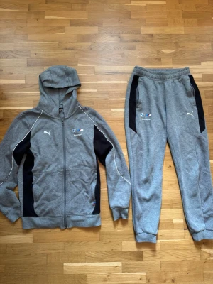 Grå BMW Puma hoodie och byxor - Säljer ett grått set med hoodie och byxor från Puma i samarbete med BMW Motorsport. Hoodien har dragkedja, huva och svarta detaljer längs sidorna samt broderade loggor på bröstet. Byxorna har resår i midjan och muddar vid benslut. Materialet är mjukt och bekvämt, perfekt för chill.