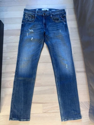Replay Anbass slim fit - Säljer ett par blå jeans från Replay med skinny fit och slitna detaljer framtill. Jeansen har klassisk femficksdesign, dragkedjegylf och Replay-lapp bak i midjan. Materialet är denim i bomull med stretch för extra komfort.