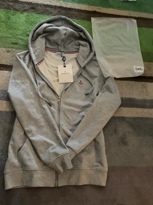 Grå zip hoodie från Moncler - Snygg grå hoodie från Moncler med dragkedja framtill och huva med dragsko. Klassisk Moncler-logga på bröstet och diskreta röda och blå detaljer vid kragen. Tillverkad i mjukt bomullsmaterial som känns skönt mot huden. Perfekt för en avslappnad och stilren look. Kan gå ner i pris vid snabb affär. Taggen var av när jag fick den! Den har inte använts!