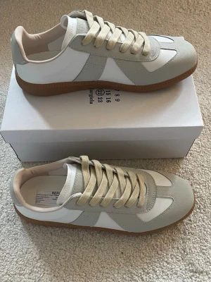 Maison Margiela  sneakers - Snygga Maison Margiela  sneakers i vitt och ljusgrått med beige detaljer. Ovandelen är i läder och mocka, och sulan är i gummi med klassisk brun färg. Perfekta för dig som gillar minimalistisk och stilren design.