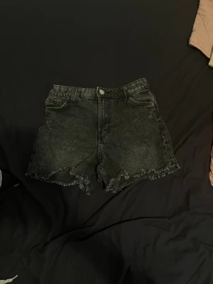 Svarta denimshorts med fransig kant - Säljer ett par svarta denimshorts med fransig rå kant och klassisk femficksmodell. Shortsen har hög midja och stängs med knapp och dragkedja. Perfekta till sommaren och ger en cool, avslappnad vibe.