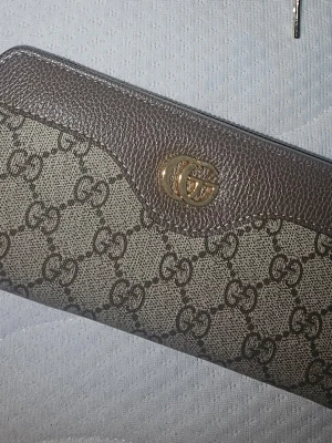 Supreme plånbok brun - Snygg plånbok i klassiskt GG Supreme-mönster. Plånboken är beige och brun med detaljer i brunt läder och har ett guldfärgat GG-logo framtill. Perfekt för dig som gillar lyxiga accessoarer och tidlös design.