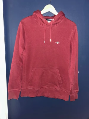 Vinröd hoodie från GANT - Säljer en röd hoodie från GANT med liten logga på bröstet. Hoodien har huva med magficka. Perfekt för dig som gillar streetwear och vill ha en enkel men snygg look.