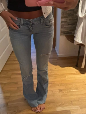 Ljusblå bootcut jeans i tall  med slitningar - Säljer ett par ljusblå bootcut jeans med snygga slitningar på låret och knät. Jeansen har klassisk femficksdesign, låg midja och broderade detaljer på bakfickorna. Perfekta för en avslappnad och trendig look. Jeansen är i tall modell.