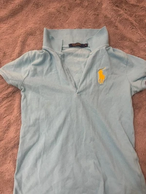Ljusblå pikétopp från Polo Ralph Lauren - Säljer en ljusblå pikétopp från Polo Ralph Lauren med klassisk krage och gul broderad logga på bröstet. Toppen har korta ärmar och är perfekt för en sportig och stilren look.